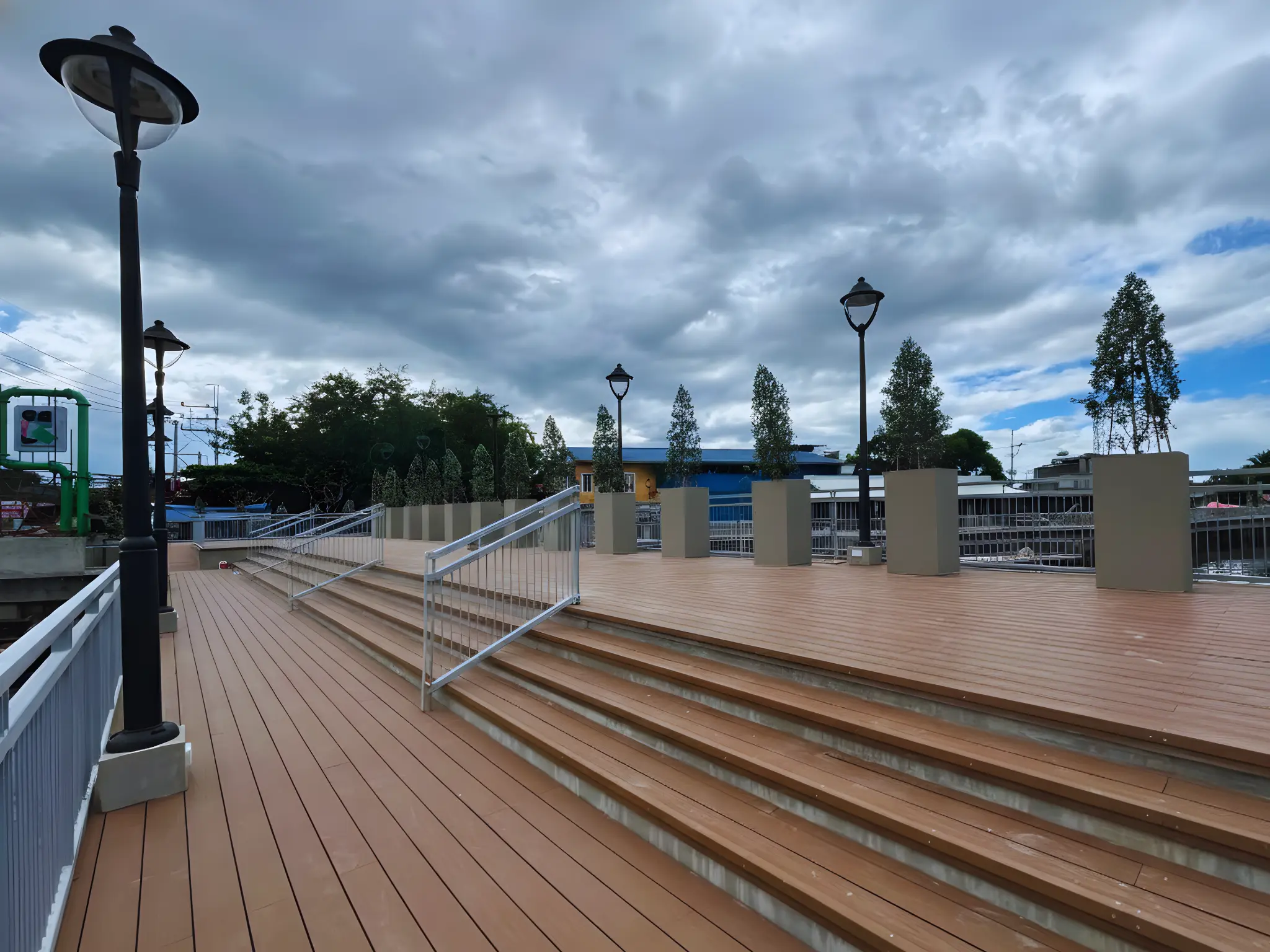 A wide public walkway or pier made of brown wood-textured planks. The area features wide wooden steps, metal handrails, and tall black street lamps. The background shows a cloudy sky and distant greenery.