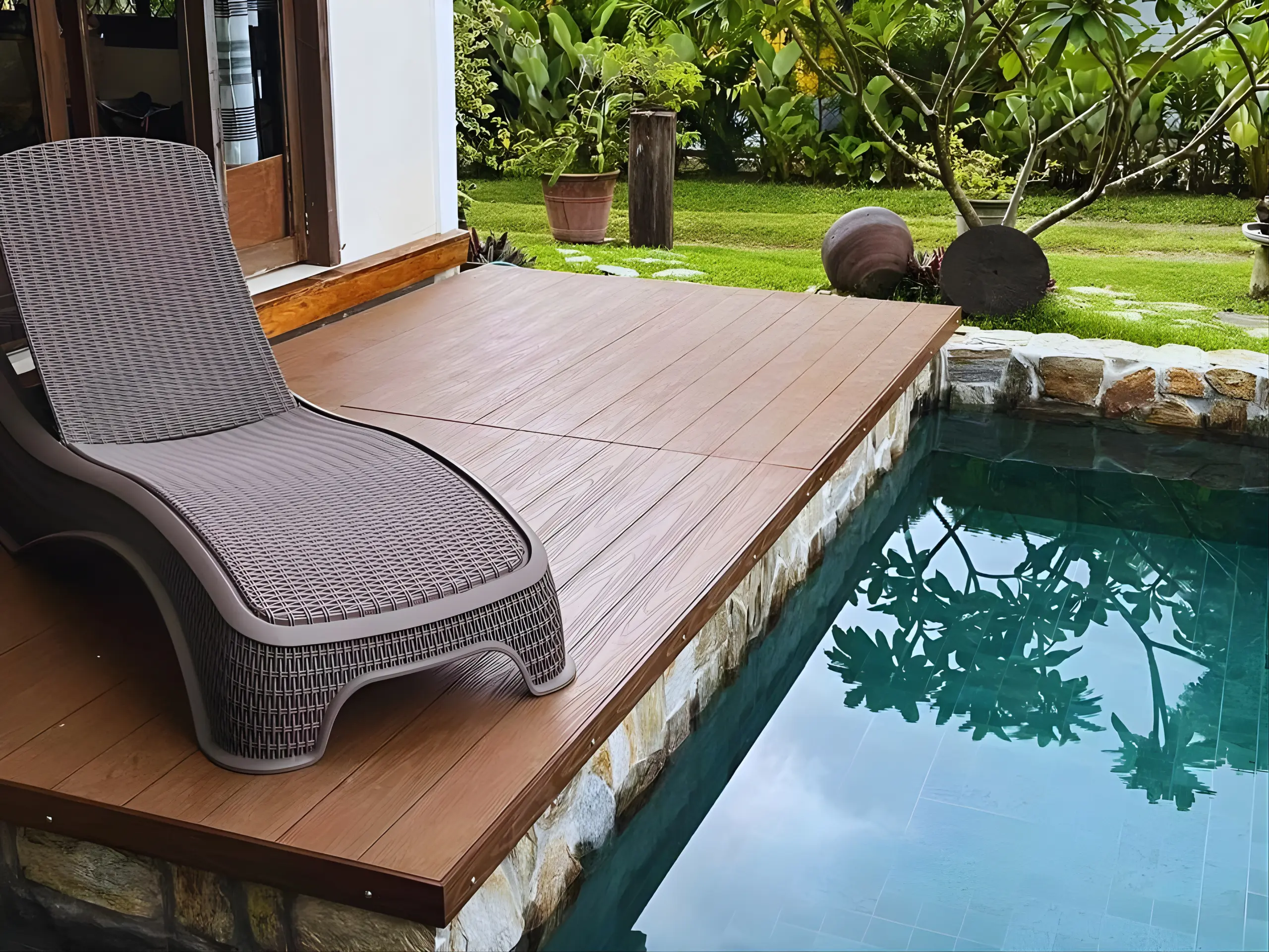 A close-up of a grey wicker lounge chair sitting on a brown wood deck. The deck is built right up to the edge of a clear blue swimming pool with stone tiling. A lush green garden with various plants and trees is visible in the background.